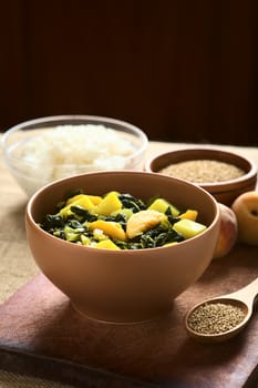 Bowl of spinach, peach and potato curry dish with sesame seeds and cooked rice in the back photographed with natural light (Selective Focus, Focus in the front of the curry dish)