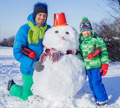 Happy liitle boy with his father building snowman outside in winter time