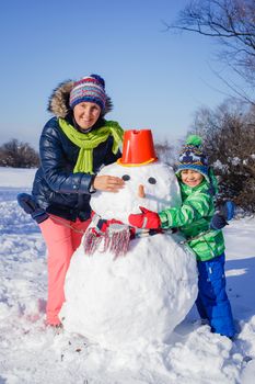 Happy liitle boy with his mother building snowman outside in winter time