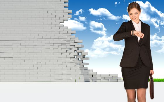 Businesswoman holding suitcase and looking at watch. Dilapidated brick wall, green meadow and sky as backdrop. Business concept