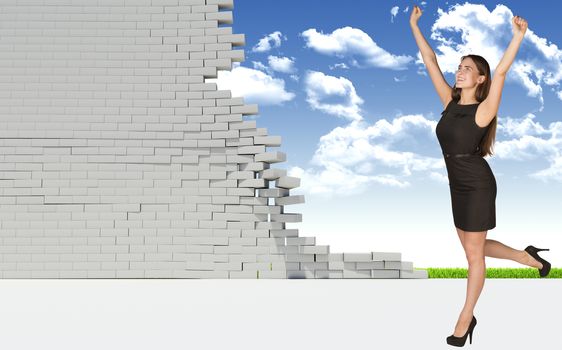 Beautiful businesswoman in dress happily raised his hands up, looking at camera. Dilapidated brick wall, green meadow and sky as backdrop. Concept of freedom