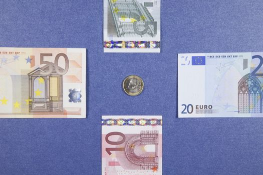One Euro coin between five ten twenty fifty banknotes on blue background