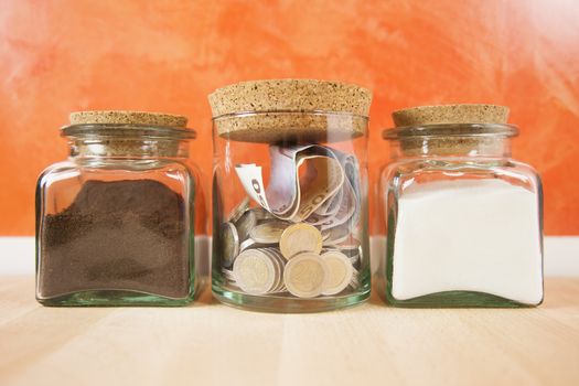 Euro coins and banknotes into crystal jar closed with cork plugs between coffee and white sugar on orange background