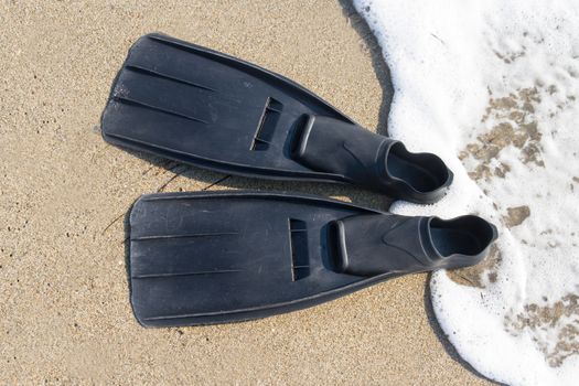 Black plastic flippers on sea sand and wavy sea with foam.