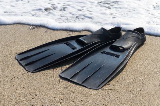 Black plastic flippers on sea sand and wavy sea with foam.