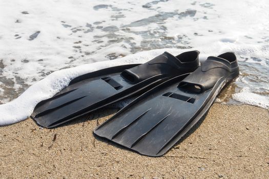 Black plastic flippers on sea sand and wavy sea with foam.