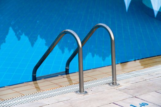 Close up of stair at blue swimming pool with reflection of trees.
