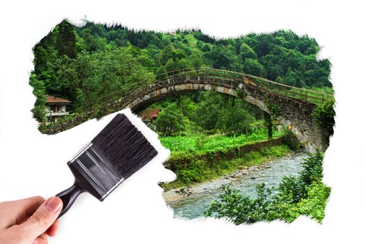 Male hand holding paint brush and painting plateau picture with bridge inside.