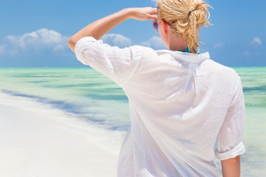 Happy woman enjoying, relaxing joyfully in summer by tropical blue water. Beautiful caucasian model  wearing white beach tunic on vacations looking down picture perfect Paje beach, Zanzibar, Tanzania.