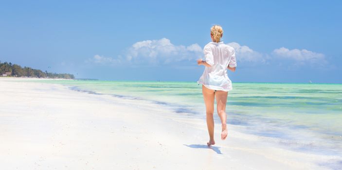 Happy woman having fun, enjoying summer, running joyfully on tropical beach. Beautiful caucasian model  wearing white beach tunic on vacations on picture perfect Paje beach, Zanzibar, Tanzania.