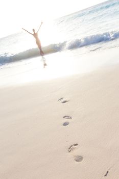 Woman, arms raised, enjoying summer on tropical beach in in sunset. Beautiful caucasian model wearing bikini on vacations on sandy beach. Footprints in sand. 