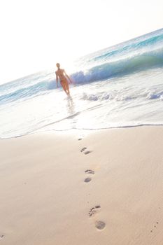 Happy woman enjoying in summer, running joyfully on tropical beach in sunset. Beautiful caucasian model wearing bikini on vacations on sandy beach. Footprints in sand. 