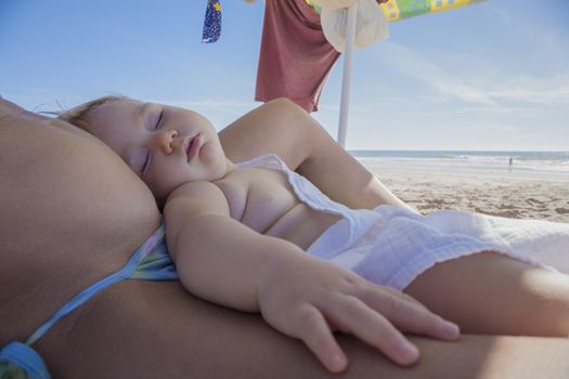 one year baby with white cloth sleeping on mom tummy at beach under parasol