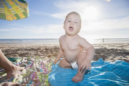 one year baby white diaper sit looking at camera backlighting over cyan towel at beach next to Conil Cadiz Spain