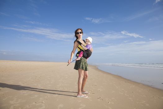 mother with green miniskirt black sunglasses carrying her one year baby with white hat in front rucksack walking at a beach