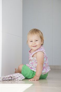 happy blonde one year baby green shorts smiling sitting on grey white floor