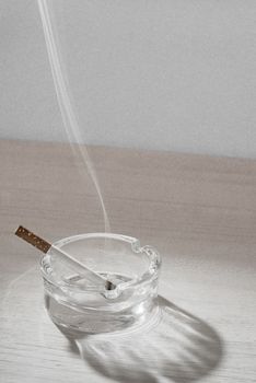 A reasently lid sigarette rests in a glass ashtray while the smoke floats up in the air.