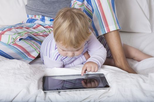 eighteen month aged blonde baby with brunette woman mother reading digital tablet on white bed