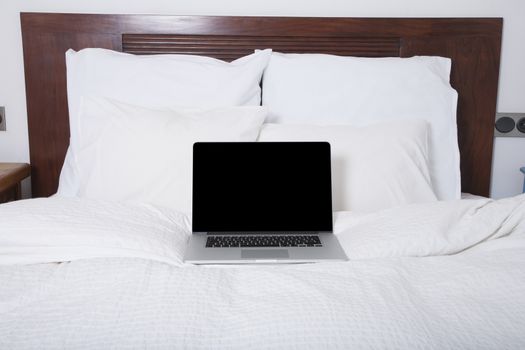 blank screen grey laptop open keyboard on white bed with pillow and cushions wood headboard