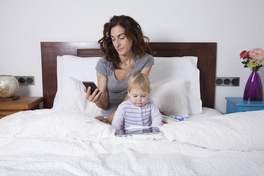 eighteen month aged blonde baby watching digital tablet and brunette woman mother at smartphone on white bed