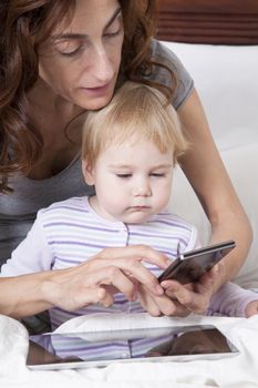 eighteen month aged blonde baby and brunette woman mother watching smartphone and digital tablet on white bed