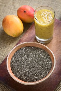 Chia seeds (lat. Salvia hispanica) in clay bowl with mango and chia juice in the back photographed with natural light. Chia seeds are considered a superfood containing proteins, omega fats, minerals and antioxidants. (Selective Focus, Focus in the middle of the bowl and the front rim of the glass)