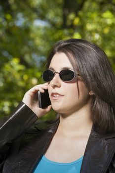 pregnant young woman with blue shirt and brown leather jacket calling phone over green trees background street
