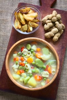 Overhead shot of Bolivian traditional Sopa de Mani (peanut soup) made of meat, pasta, vegetables (pea, carrot, potato, broad bean, pepper, corn) with ground peanut in wooden bowl, traditionally served with fried potatoes, photographed with natural light (Selective Focus, Focus on the soup)