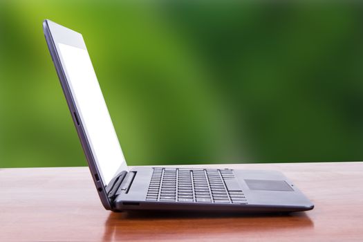 Tablet laptop with white blank screen on wooden table, side view, green natural background.