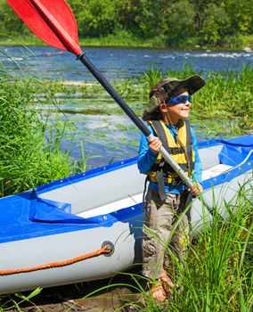 Portrait of happy cute boy holding paddle near a kayak on the river, enjoying a lovely summer day