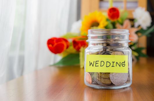 Many world coins in a money jar with wedding label on jar, beautiful flowers on background