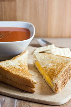 A grilled cheese sandwich with a bowl of tomato soup and saltine crackers on a wooden serving board.