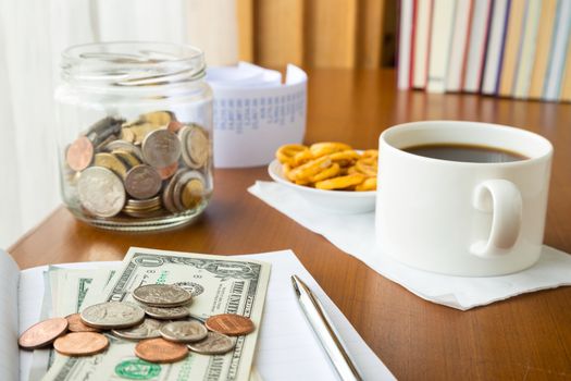 Many coins and banknotes place on notebook with a money saving jar, bills and coffee break on table, home finance concept