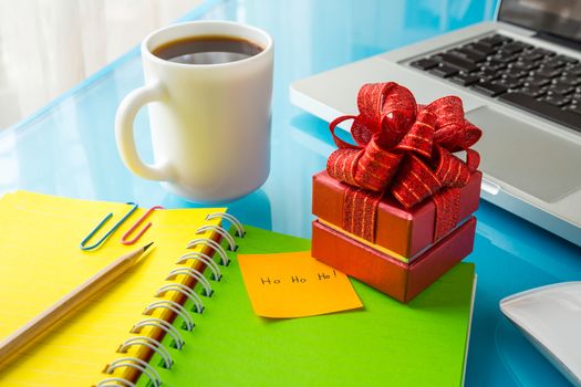 Red present box and paper note with Merry christmas message: Ho Ho Ho! place on office table with laptop, colorful diary and a cup of coffee, concept to christmas gift and holiday season