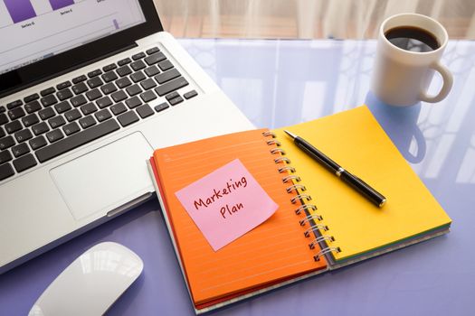 Message on paper with Marketing Plan word on note stick on colorful book with laptop and a cup of coffee on glass table, top view image