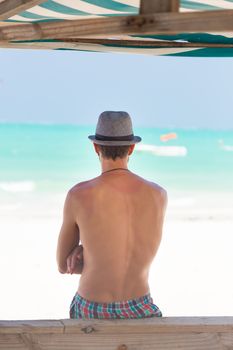 Man hiding in shade from burning tropical sun on picture perfect tropical sandy beach. Summer leisure. Shot from back.