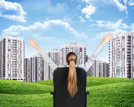 Businesswoman sitting on office chair with her hands outstretched. Green hills with buildings and sky as backdrop