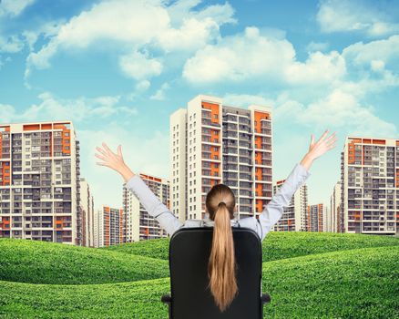 Businesswoman sitting on office chair with her hands outstretched. Green hills with buildings and sky as backdrop