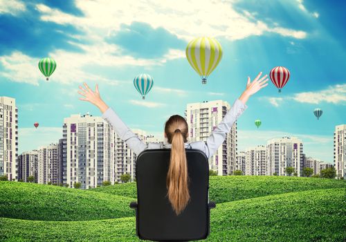 Businesswoman sitting on office chair with her hands outstretched. Green hills with buildings and sky as backdrop