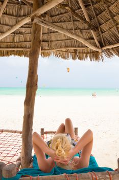 Women sunbathing on dack chair under wooden umbrella on stunning tropical beach. Kiteboarders turquoise blue lagoon of Paje beach, Zanzibar, Tanzania in the background.