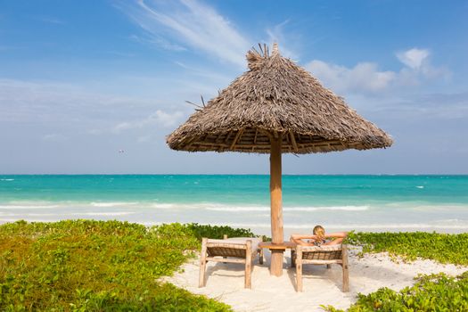 Women sunbathing on dack chair under wooden umbrella on stunning tropical beach. Turquoise blue lagoon of Paje beach, Zanzibar, Tanzania in the background.