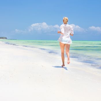 Happy woman having fun, enjoying summer, running joyfully on tropical beach. Beautiful caucasian model  wearing white beach tunic on vacations on picture perfect Paje beach, Zanzibar, Tanzania.