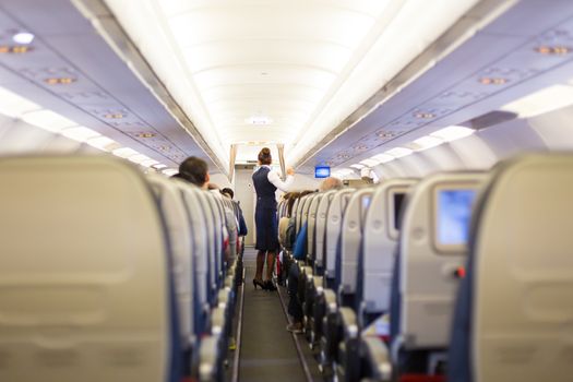 Interior of airplane with passengers on seats and stewardess in uniform walking the aisle. 