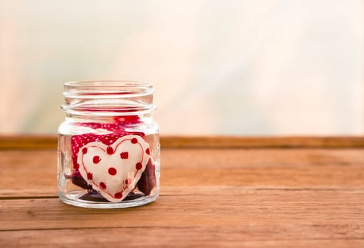 Group of cute red tone hearts handmade crafts from cotton and silk cloth in glass jar place on wood surface with white curtain on background, love and valentine's day symbol