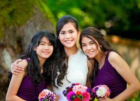 Biracial bride standing with her two bridesmaids outside, smiling and holding flower bouquet 