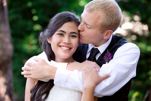 Caucasian groom lovingly kissing his biracial bride on cheek. Diverse couple
