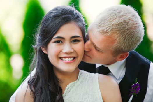 Caucasian groom lovingly kissing his biracial bride on cheek. Diverse couple