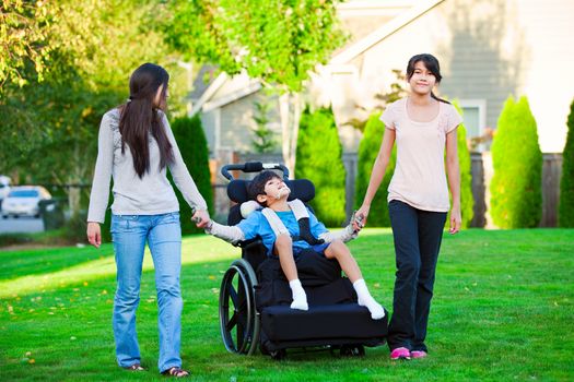 Disabled little boy in wheelchair walking with sisters on glassy lawn