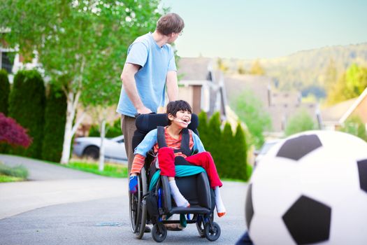 Caucasian father helping disabled biracial son in wheelchair play soccer outdoors on street