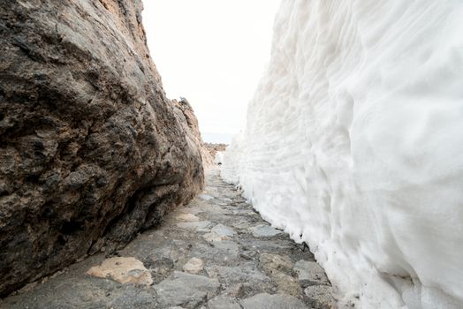 Foot path in mountains between rock wall and snow